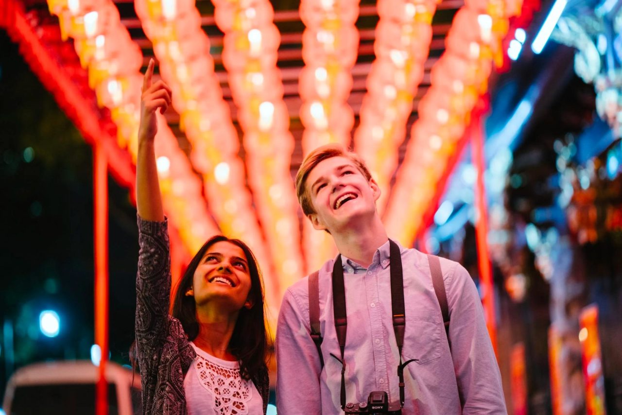 dos turistas sonríen y miran las luces que se muestran en un lugar público.