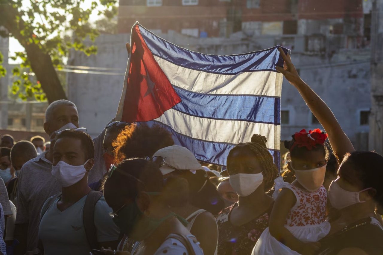 una multitud de personas sostiene una bandera cubana mientras se reúnen afuera.