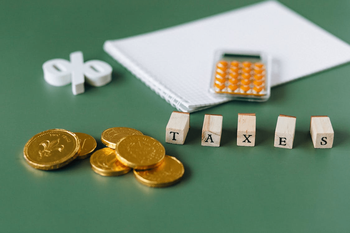 A notebook, coins, and blocks of letters spelling “Taxes” laid out on a green surface.