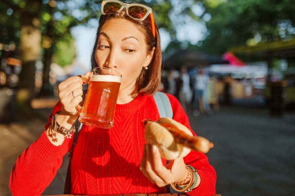 Young happy asian traveler woman drinks mug of beer with hotdog in Germany, beer and food festival concept