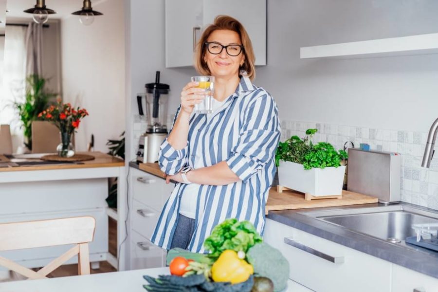 Una mujer con gafas está parada en una cocina, sosteniendo un vaso de agua.