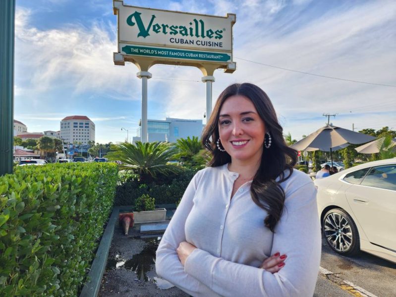 Nicole Valls con camisa blanca sonriendo frente a un cartel de cocina cubana de Versalles