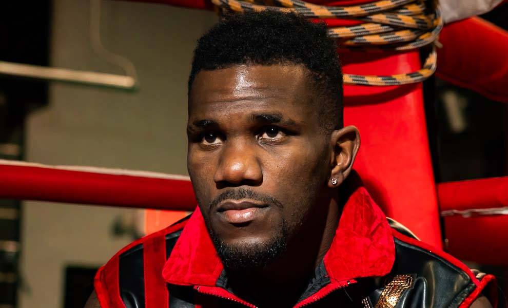 Un hombre con una chaqueta roja y negra sentado en un ring de boxeo, listo para un combate.