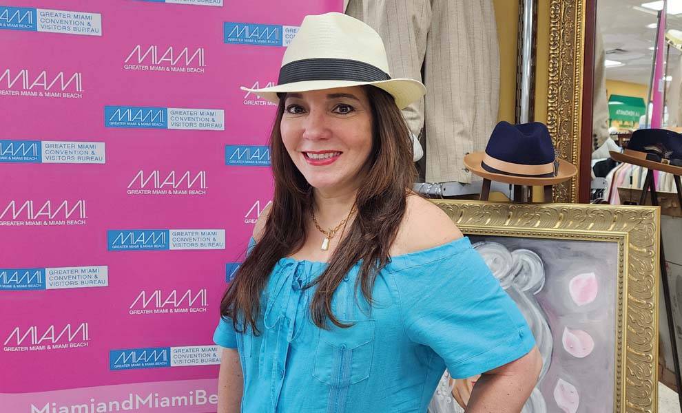 Mujer con sombrero y camisa azul parada frente a una pared rosa.