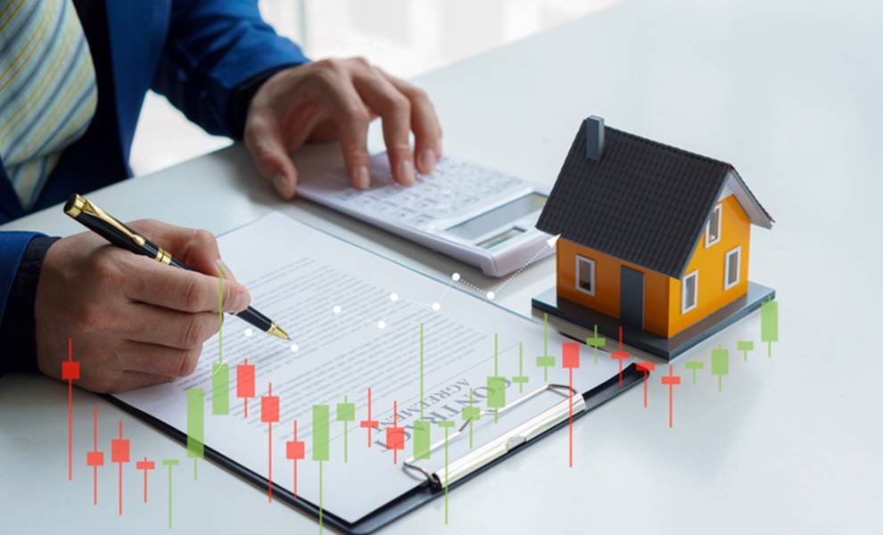 Un hombre escribiendo en papel con un modelo de casa y un gráfico del mercado de valores al fondo.