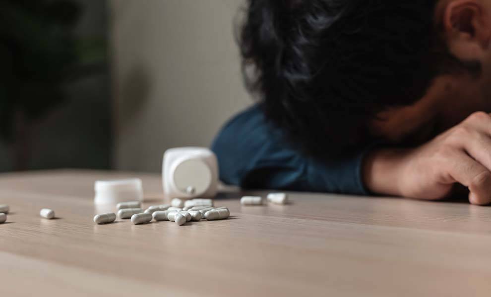Un hombre durmiendo sobre una mesa con pastillas esparcidas.