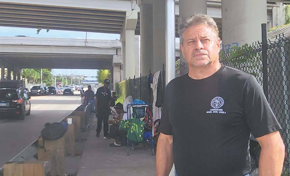A man stands beside a parked car in front of a bridge, showcasing the structure and surrounding landscape.