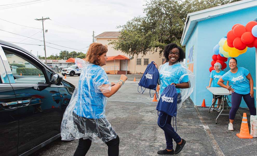 Two women in blue Hope For Miami shirts and bags stand in front of a blue building.