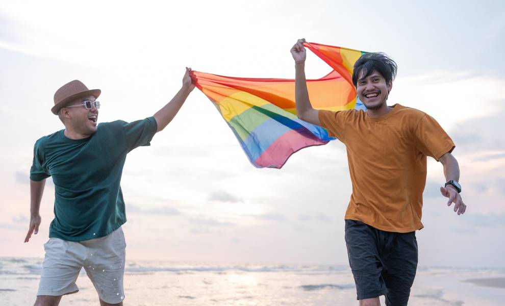 Dos hombres corren por la playa, portando orgullosamente una vibrante bandera del arcoíris, que simboliza la diversidad y la unidad.