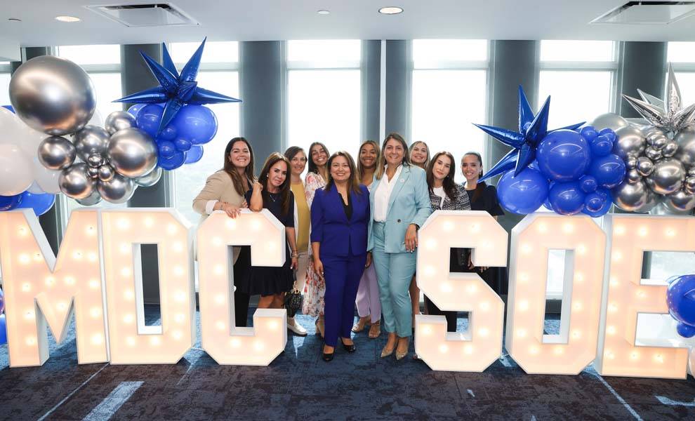 Un grupo de mujeres posa juntas frente a un gran cartel que dice "MDC SOE", mostrando unidad y colaboración.