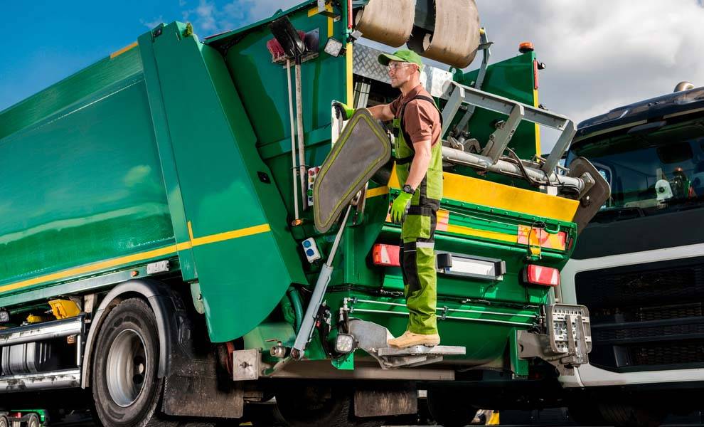 A man stands on the back of a green garbage truck.