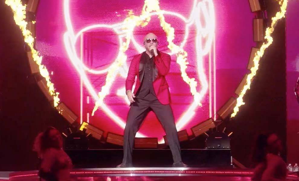 Pitbull actuando en el escenario frente a un fuego y un fondo rosa.