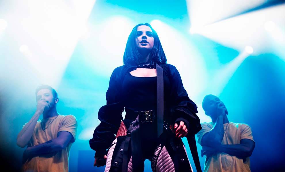Becky G in a black outfit confidently stands on stage.