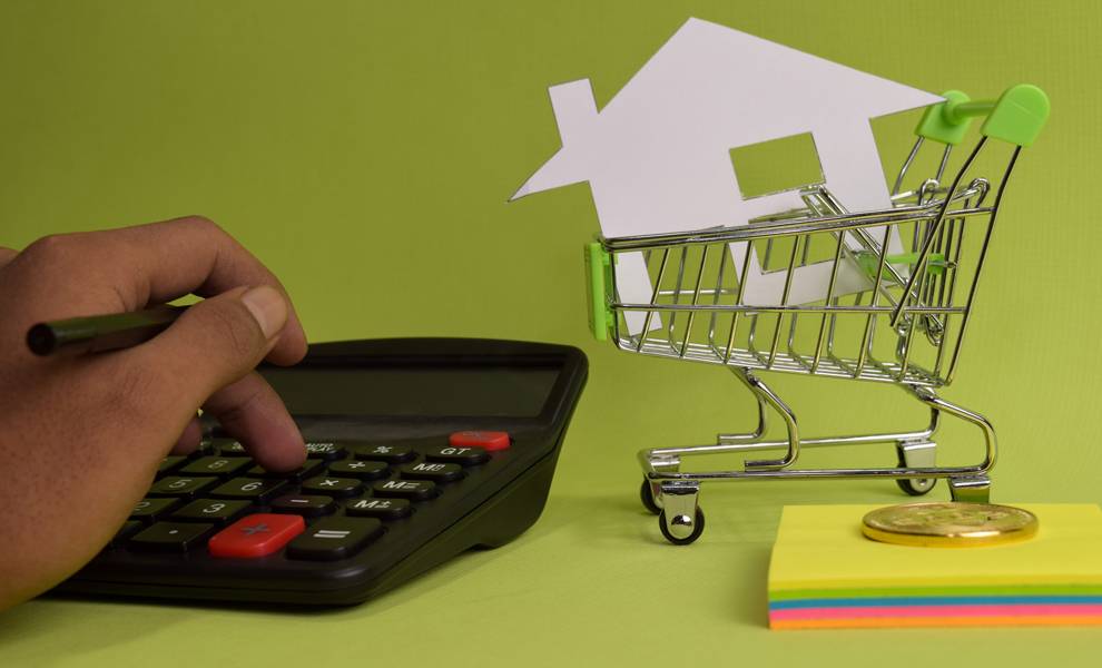 A hand holds a calculator and a house model against a green background, symbolizing shopping for home insurance.