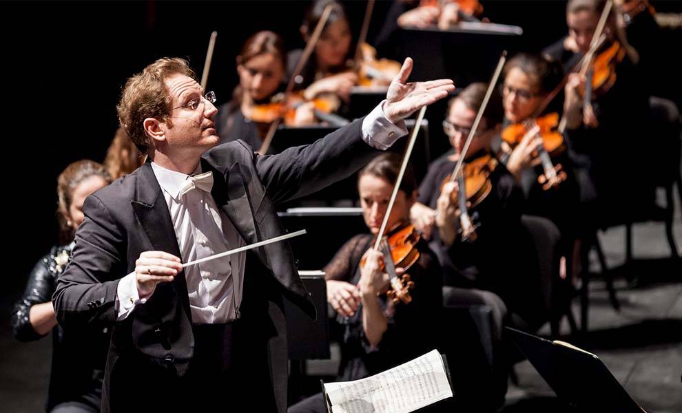 A conductor in a tuxedo leads an orchestra, surrounded by musicians playing various instruments in harmony.