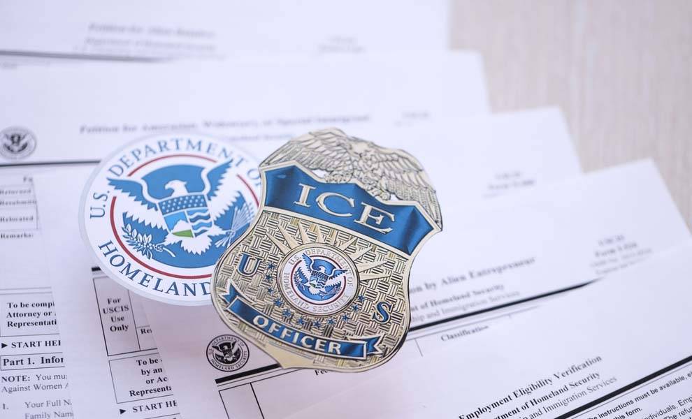 An ICE badge and passport placed atop various documents, symbolizing travel and identification.