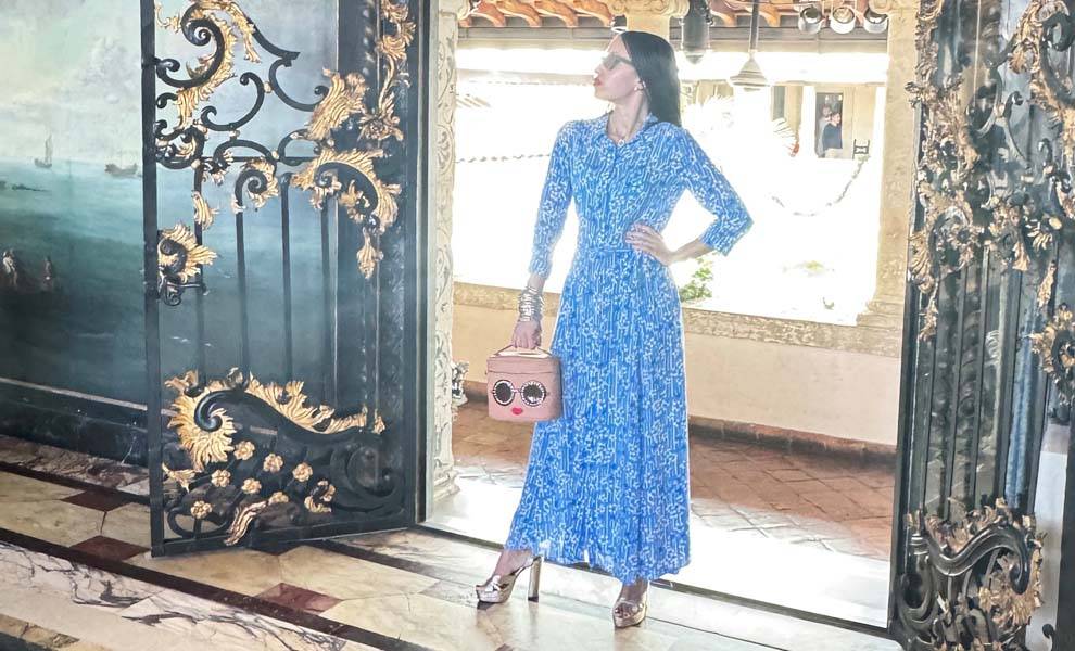 A woman in a blue dress stands gracefully in front of a door, exuding elegance and poise.