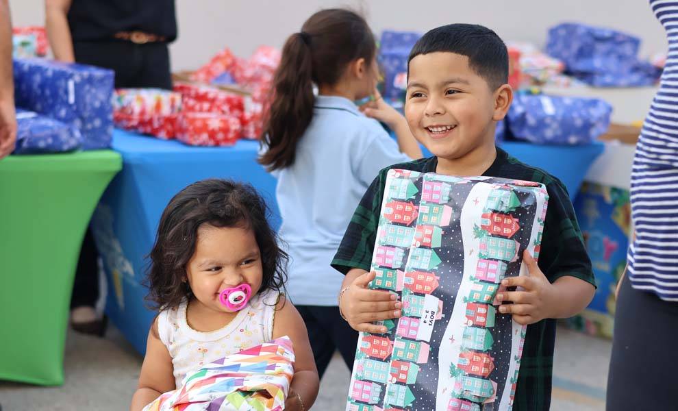 Dos niños sostienen alegremente regalos coloridos en un evento festivo, rodeados de decoraciones y un ambiente alegre.
