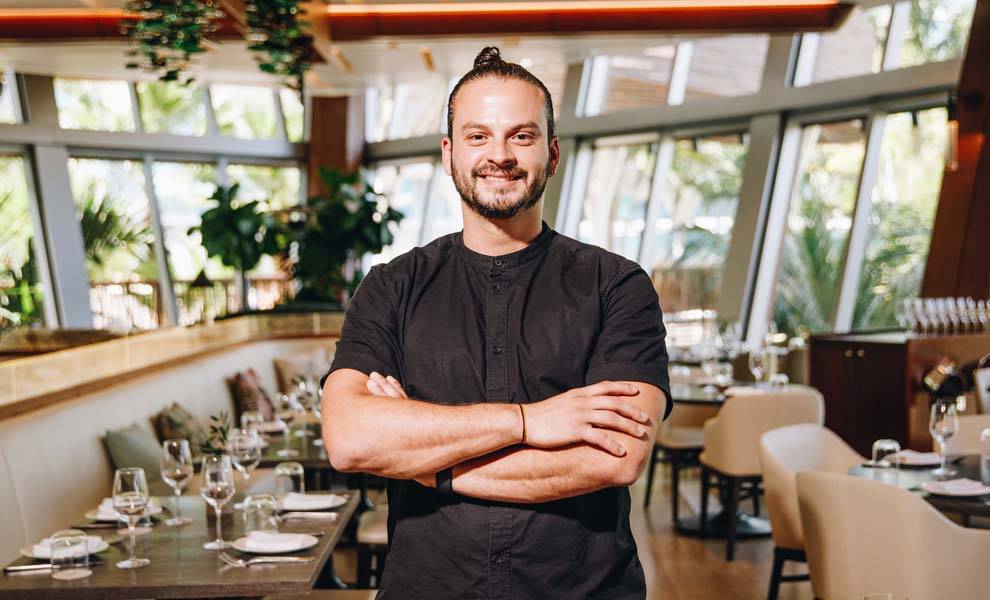 Un hombre con una camisa negra se encuentra de pie con confianza frente a un restaurante, mostrando una atmósfera acogedora.