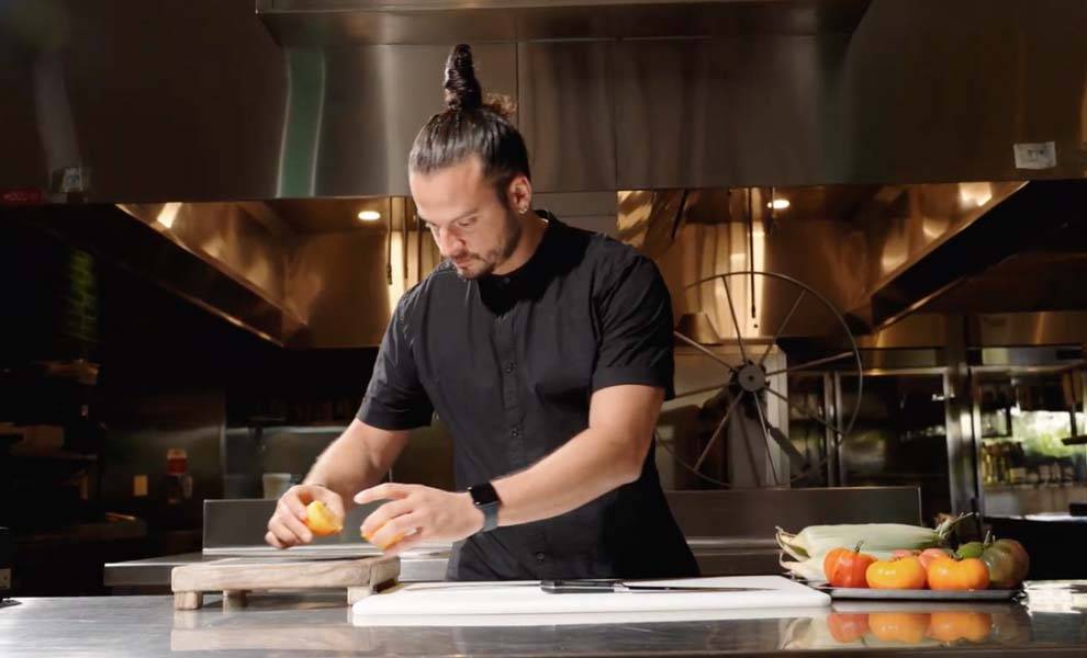 A man with a ponytail skillfully slices an orange on a cutting board, showcasing his culinary skills.