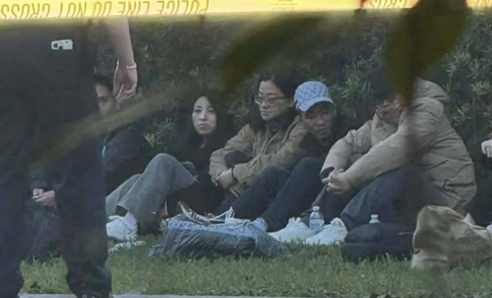 Un grupo de inmigrantes chinos sentados en el césped, con una cinta policial visible al fondo.