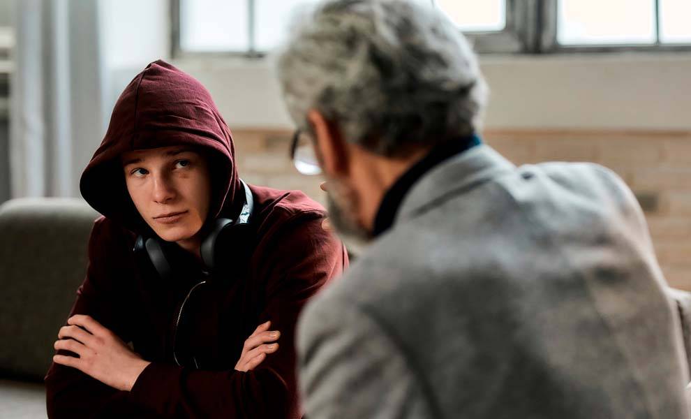 Un hombre con una sudadera con capucha se sienta al lado de otro hombre, ambos conversando en un ambiente informal.