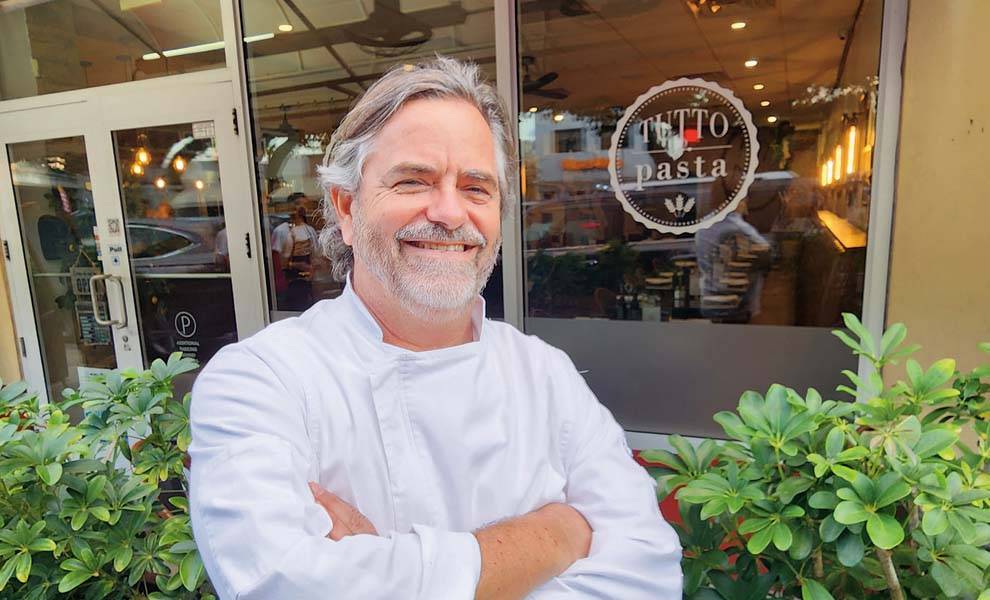 A man in a chef's uniform poses outside a Tutto Pasta, showcasing his culinary expertise.