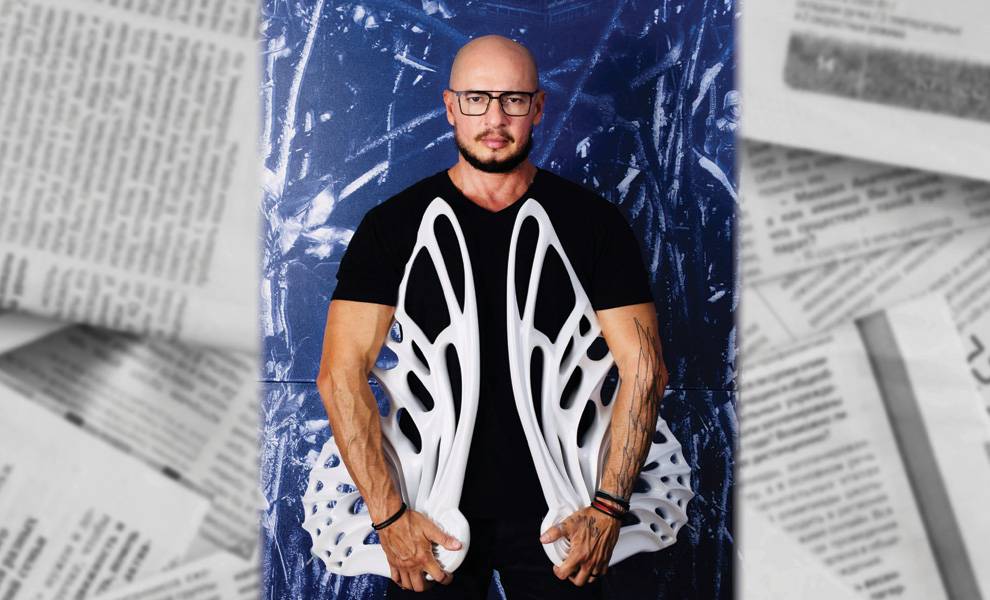 Un hombre que lleva gafas y una camisa negra sostiene un par de zapatos, sonriendo a la cámara.