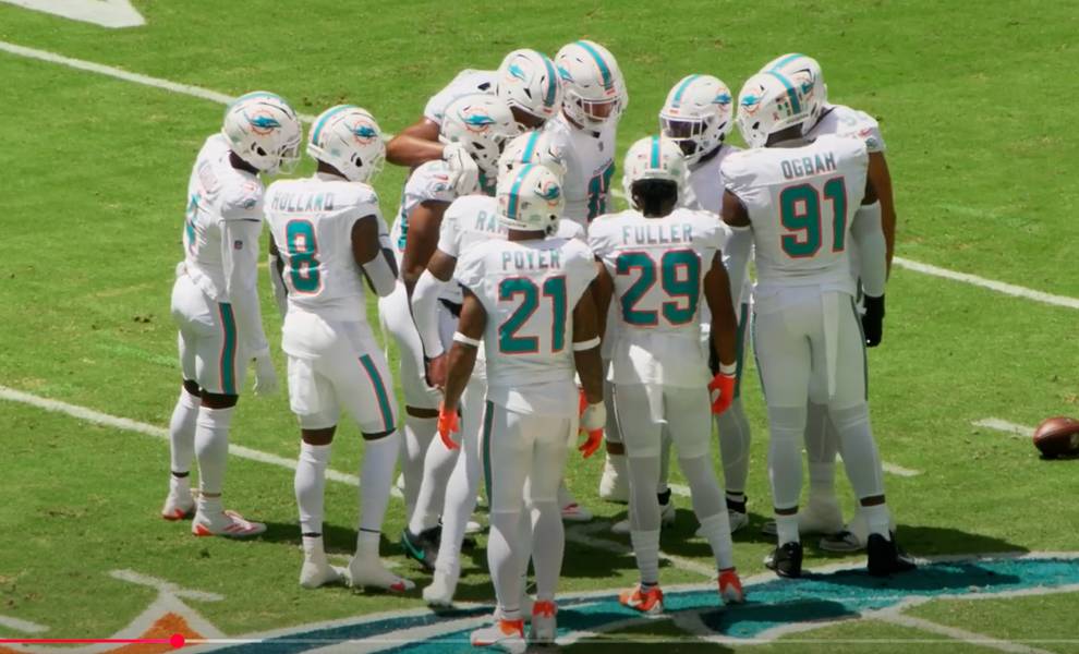 Los jugadores de fútbol de los Miami Dolphins se reunieron en el campo con uniformes blancos con detalles en aguamarina y naranja, elaborando estrategias antes de la siguiente jugada.