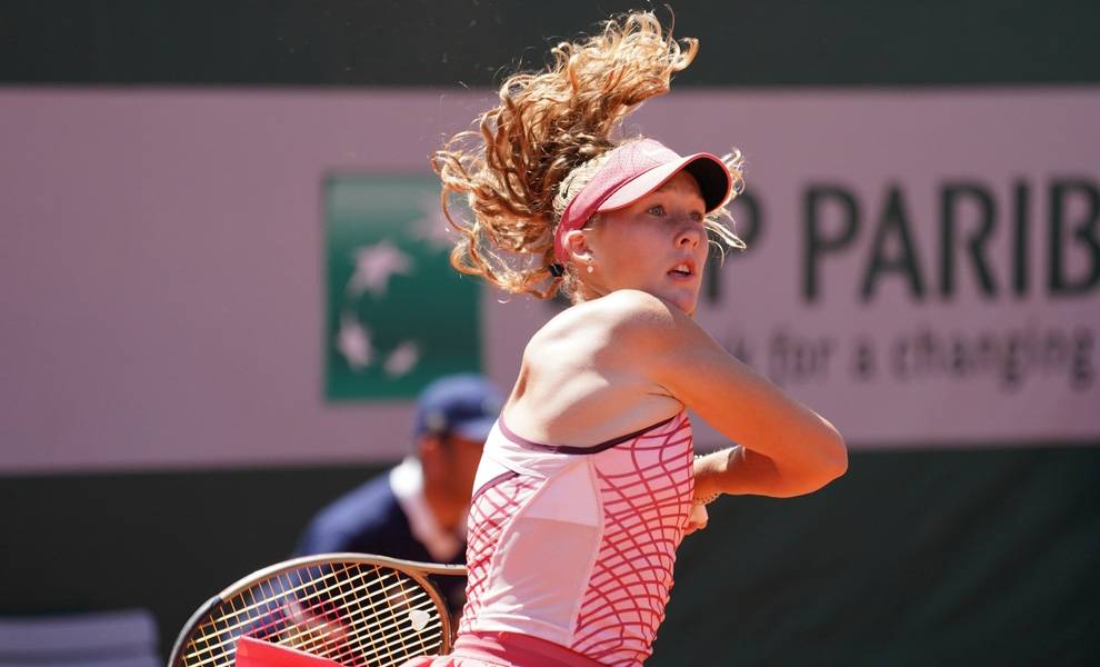 Tennis player Mirra Andreeva in athletic wear serves a tennis ball on a sunny court.