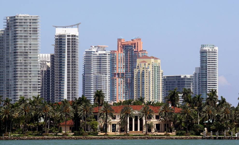 A scenic view of Miami's skyline featuring modern high-rise buildings behind a luxurious waterfront mansion surrounded by palm trees.