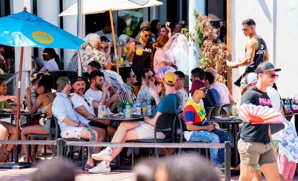 Un grupo de personas disfrutando de bebidas en mesas a la sombra de sombrillas en un animado entorno al aire libre durante las vacaciones de primavera en Miami.