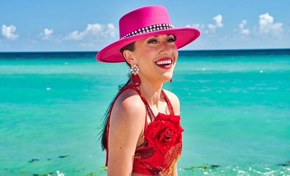 A woman wearing a red dress and a pink hat stands on the beach, with waves gently lapping at the shore behind her.