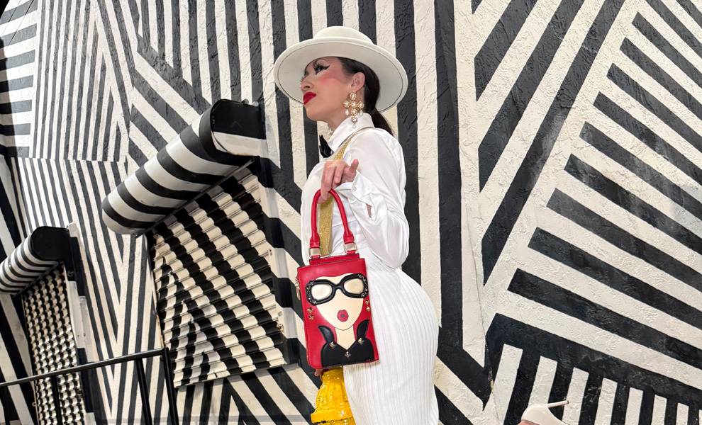 Yli Liriano poses confidently in front of a bold black-and-white geometric mural. She wears a white pinstripe outfit with a wide-brimmed hat and statement earrings, holding a red handbag featuring a playful cartoon face design.