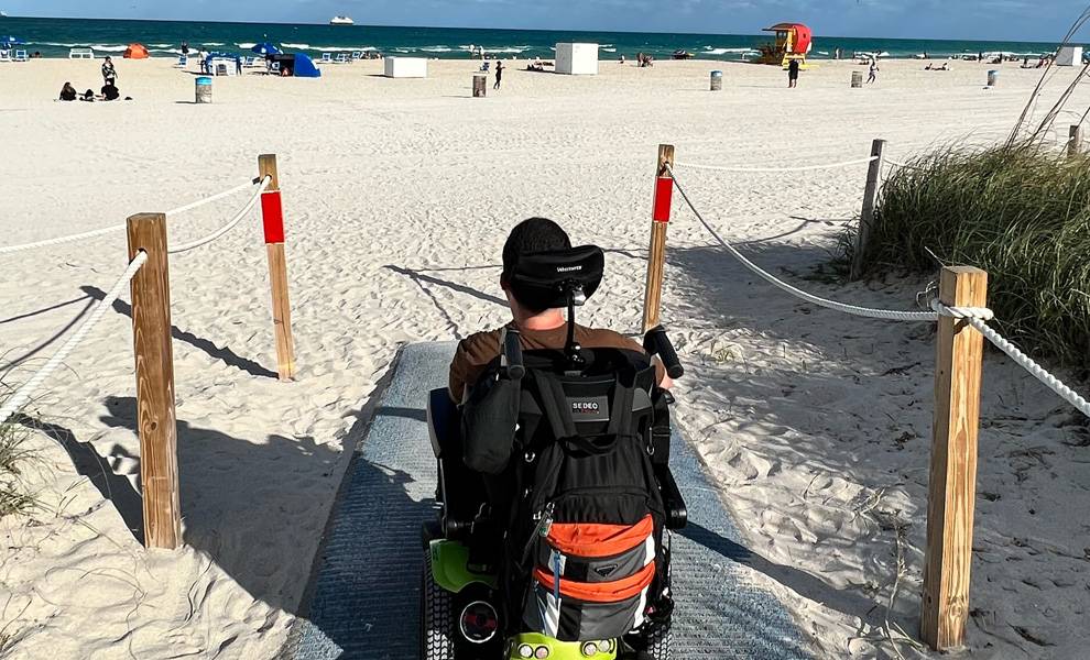 A man in a wheelchair enjoys the beach, with sand and waves visible in the background.