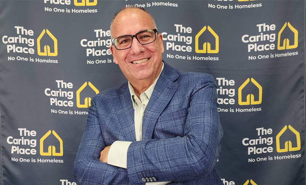 Antonio Villasuso in glasses and a blue suit stands before a wall displaying The Caring Place logo.