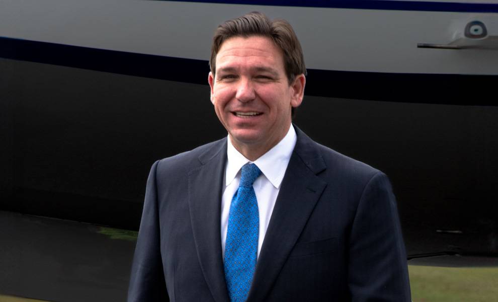 Florida Governor Ron DeSantis in a suit and tie stands confidently in front of a parked airplane on a clear day.