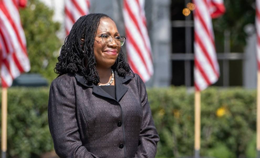 Justices Ketanji Brown Jackson in a suit and tie stands confidently in front of multiple American flags.