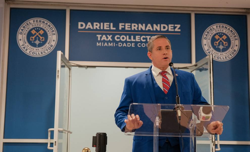 El recaudador de impuestos de Miami-Dade, Dariel Fernández, vistiendo un traje azul y corbata roja, habla con los medios de comunicación.