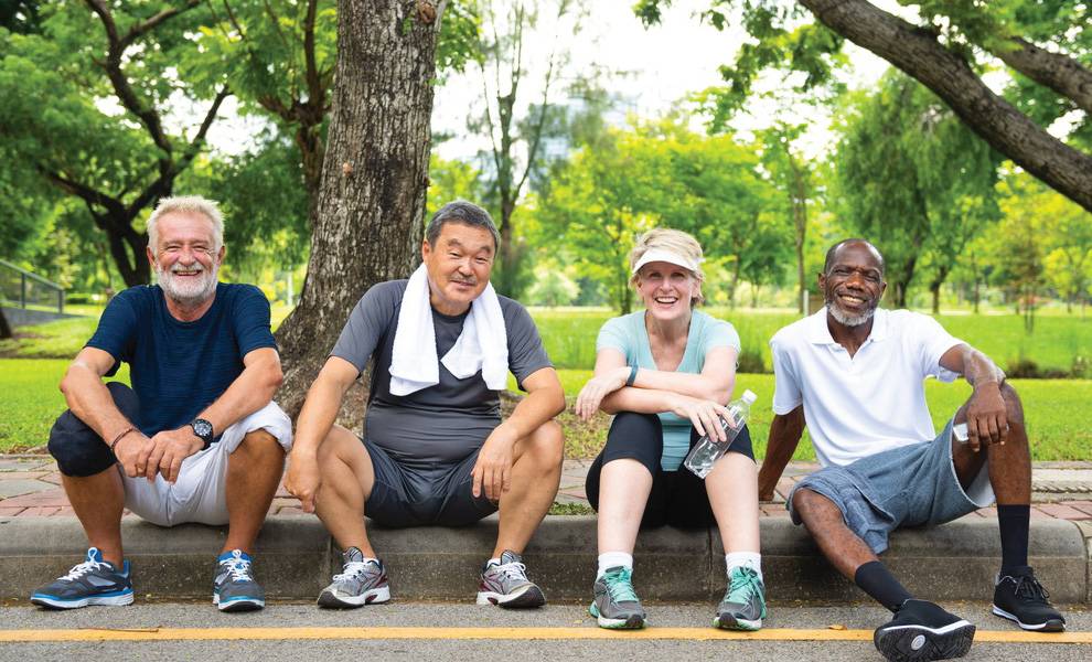 Cuatro adultos mayores de diversos orígenes se sientan en la acera de un parque verde después de hacer ejercicio, sonriendo a la cámara.