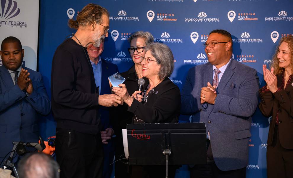 Mayor Daniella Levine Cava holding a microphone presents a small glass award to a man during a CITT event. Several people stand beside them applauding.