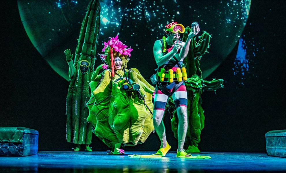 Four stage performers in bright green, plant-themed costumes pose under blue and teal theatrical lighting.