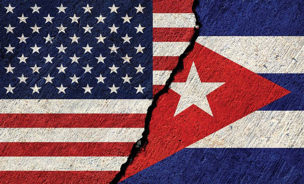 A split image of the United States and Cuban flags painted on a textured wall, divided down the center by a jagged crack, symbolizing political tension or division between the two countries.