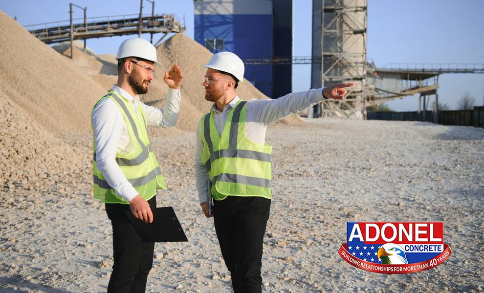 Dos trabajadores de la construcción, que visten cascos blancos y chalecos de seguridad reflectantes, se encuentran en una obra de hormigón con grandes pilas de grava y estructuras transportadoras al fondo.