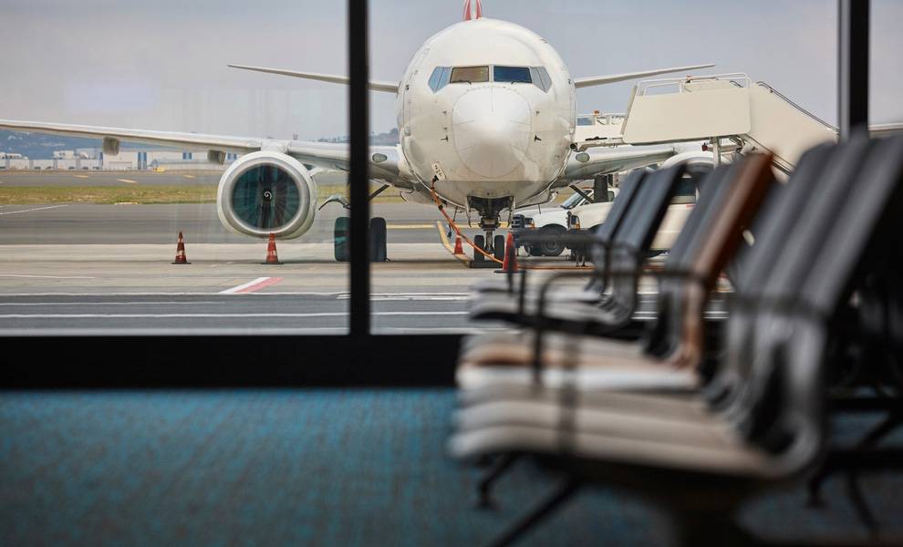 Un avión comercial está estacionado en una puerta de embarque de un aeropuerto, visto de frente a través de los grandes ventanales de la terminal.