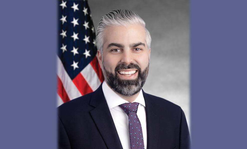 El presidente del condado de Miami-Dade, Anthony Rodríguez, sonríe en un retrato formal. Viste un traje oscuro, camisa blanca y corbata estampada, de pie frente a un fondo borroso de la bandera estadounidense.