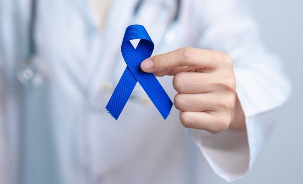 Primer plano de un profesional de la salud vestido con una bata blanca y sosteniendo una cinta azul de concienciación sobre el cáncer colorrectal entre sus dedos.