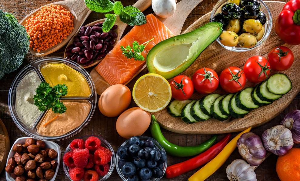 Overhead view of a wooden table filled with healthy foods including salmon, avocado, tomatoes, cucumbers, broccoli, lentils, kidney beans, eggs, olives, hummus, nuts, berries, garlic, chili peppers, and olive oil, representing a balanced weight management diet.