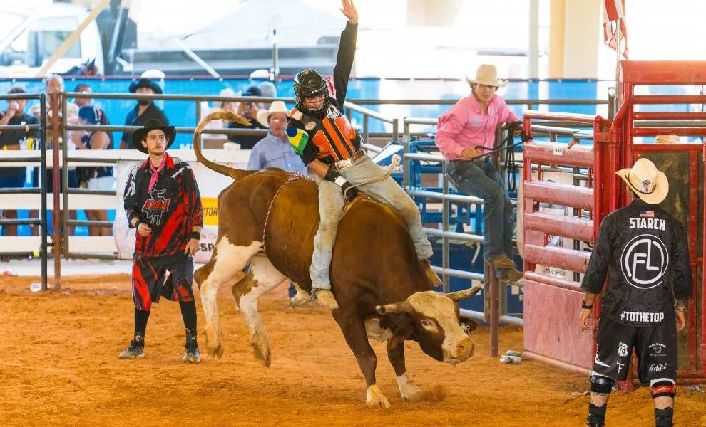 Un jinete de toros, ataviado con equipo de protección, se aferra con una mano mientras alza la otra, mientras un toro reparador levanta polvo en la arena de rodeo del CountryFest.