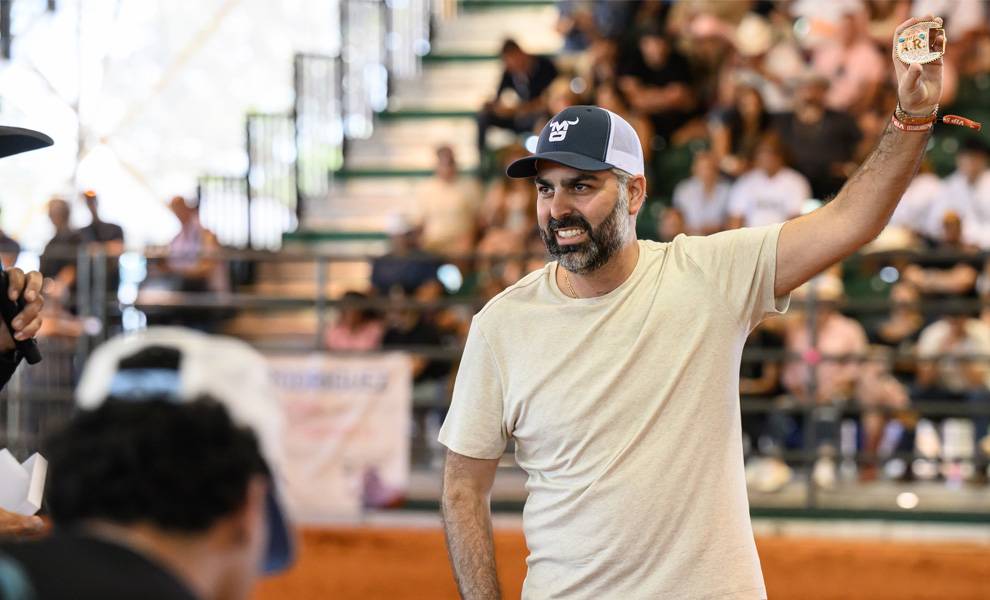 A bearded man in a baseball cap and light-colored T-shirt raises a small award or belt buckle in one hand, smiling as he stands inside an indoor arena with a seated crowd behind him at CountryFest.